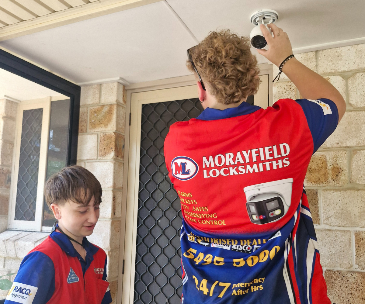 A Morayfield Locksmiths technician mounting a white security camera to a porch ceiling while an assistant watches.